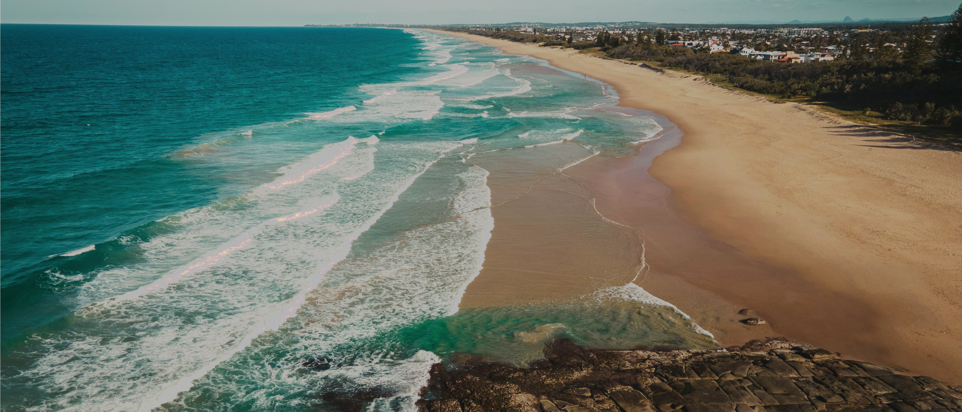 Aerial view of a Sunshine Coast beach — turquoise surf, pale sand, headland in foreground